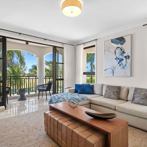 This spacious, well-lit living room has a modern vibe with a comfy sectional sofa, coffee table, and large flat-screen TV. It's decorated in neutral colors with blue accents and has a sliding glass door that opens to a balcony with pool and greenery