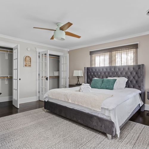 Our beautiful primary bedroom exudes a cozy atmosphere, highlighted by ample windows that invite natural light. It includes a TV, a dresser, a spacious closet, framed accents, a fan, bedside tables with lamps, and a study table with a chair.