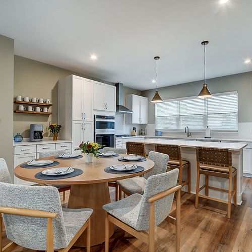 This open-concept kitchen and dining area features a round wooden table set for six. The kitchen boasts white cabinetry, stainless steel appliances, a large island with bar seating, and pendant lighting creating a welcoming space.