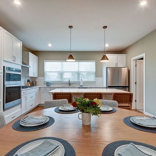 This open-concept kitchen and dining area features a round wooden table set for six. The kitchen boasts white cabinetry, stainless steel appliances, a large island with bar seating, and pendant lighting, creating a welcome space.