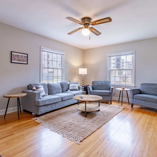 A cozy living room featuring a plush couch with soft throw pillows, two stylish accent chairs arranged for conversation, a sleek coffee table at the center, and a mounted TV above for entertainment. The room is well-lit with natural light pouring in.