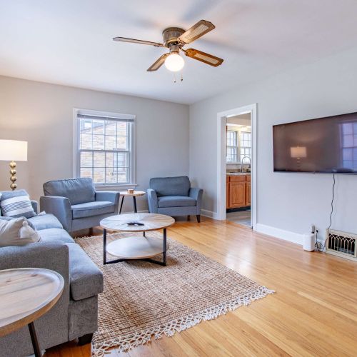 A cozy living room featuring a plush couch with soft throw pillows, two stylish accent chairs arranged for conversation, a sleek coffee table at the center, and a mounted TV above for entertainment. The room is well-lit with natural light pouring in.