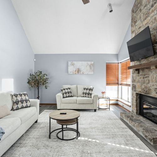 The main living area features two couches, a flat screen TV, electric fireplace, a bookshelf with books, and plenty of light!