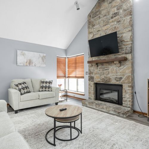 The main living area features two couches, a flat screen TV, electric fireplace, a bookshelf with books, and plenty of light!