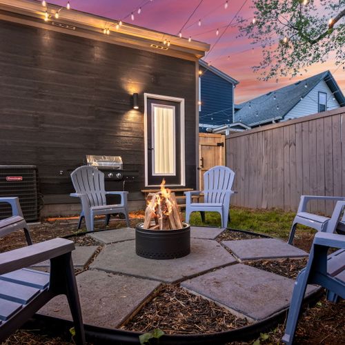 Kick back in the backyard by the modern stainless steel fire pit, blue chairs, games, and grill—outdoor vibes done right.