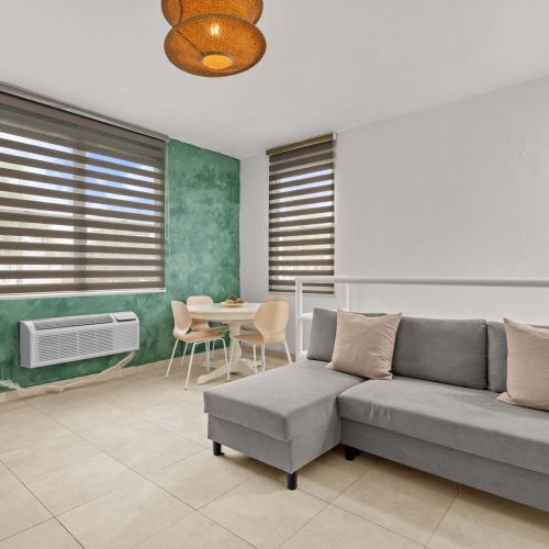 Espacio de concepto abierto. En primer plano, sofá seccional gris junto a una barandilla blanca. Al fondo, mesa de comedor redonda blanca frente a una pared verde. Dos ventanas con persianas y unidad de aire acondicionado instalada en la pared.