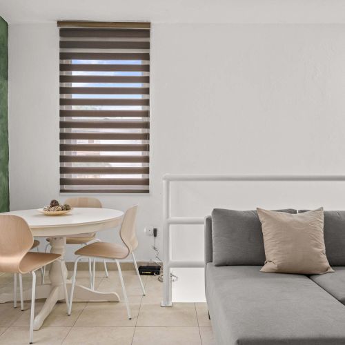 Espacio de concepto abierto. En primer plano, sofá seccional gris junto a una barandilla blanca. Al fondo, mesa de comedor redonda blanca frente a una pared verde. Dos ventanas con persianas y unidad de aire acondicionado instalada en la pared.