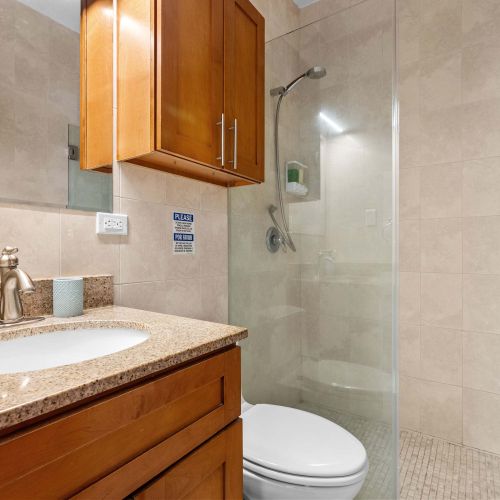 Baño con azulejos beige. A la izquierda, lavabo sobre encimera de granito y gabinete de madera suspendido. A la derecha, ducha a ras de suelo con mampara de vidrio transparente fija y cabezal de ducha cromado en la pared.