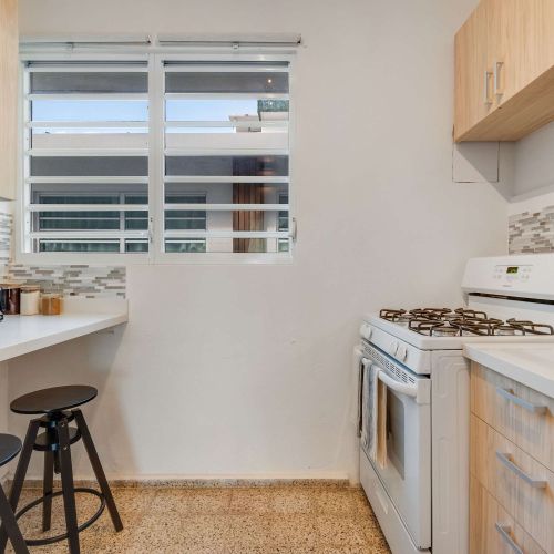 A narrow galley kitchen with light wood cabinets, a stainless steel gas stove, oven, and refrigerator. White countertops line both sides, with framed artwork on the wall and a wooden runner rug on the floor.