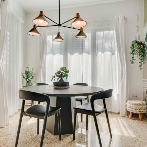 A dining area with a round black table and four matching chairs. Pendant lights hang above the table, and sheer white curtains cover the windows. Potted plants decorate the corners of the room.