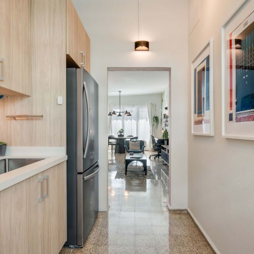 A narrow galley kitchen with light wood cabinets, a stainless steel gas stove, oven, and refrigerator. White countertops line both sides, with framed artwork on the wall and a wooden runner rug on the floor.