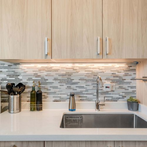 A narrow galley kitchen with light wood cabinets, a stainless steel gas stove, oven, and refrigerator. White countertops line both sides, with framed artwork on the wall and a wooden runner rug on the floor.