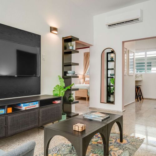 A living room with a dark sofa facing a wall-mounted TV. A small coffee table sits in front of the sofa, and open shelving with decorative items lines the wall. Neutral tones and natural light fill the space.