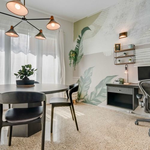 A dining area with a round black table and four matching chairs. Pendant lights hang above the table, and sheer white curtains cover the windows. Potted plants decorate the corners of the room. PC, desk and office chair