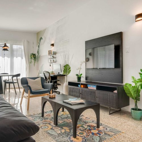 A living room with a dark sofa facing a wall-mounted TV. A small coffee table sits in front of the sofa, and open shelving with decorative items lines the wall. Neutral tones and natural light fill the space.