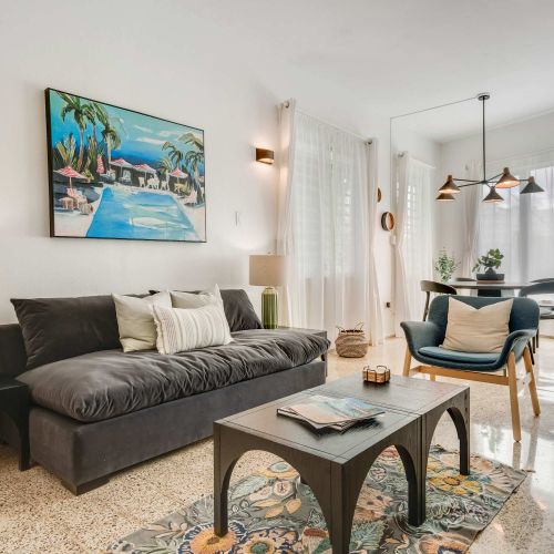 A living room with a dark sofa facing a wall-mounted TV. A small coffee table sits in front of the sofa, and open shelving with decorative items lines the wall. Neutral tones and natural light fill the space.