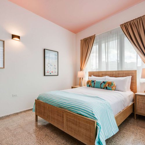 A bedroom with a queen-size bed dressed in white linens and a light blue throw. Two windows with beige curtains allow natural light. A wooden nightstand and soft lighting frame the bed.