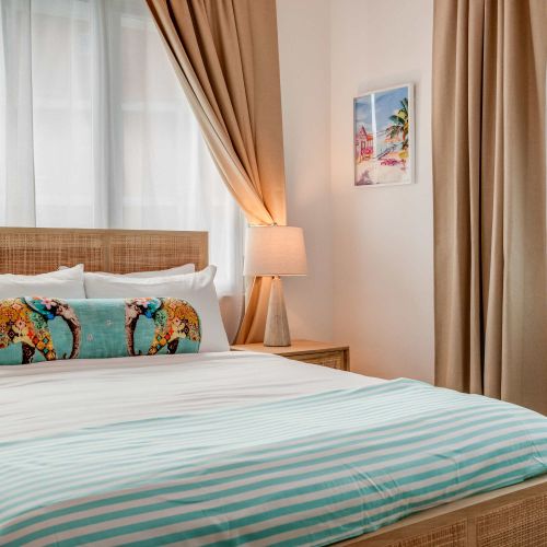 A bedroom with a queen-size bed dressed in white linens and a light blue throw. Two windows with beige curtains allow natural light. A wooden nightstand and soft lighting frame the bed.