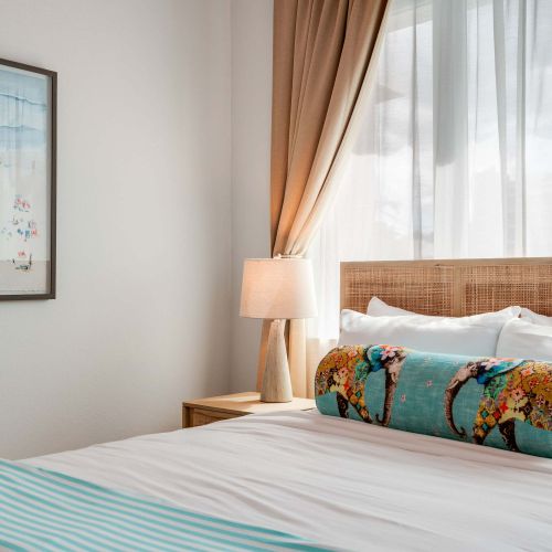 A bedroom with a queen-size bed dressed in white linens and a light blue throw. Two windows with beige curtains allow natural light. A wooden nightstand and soft lighting frame the bed.