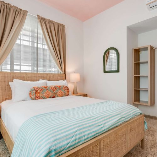 A bedroom with a queen-size bed dressed in white linens and a light blue throw. Two windows with beige curtains allow natural light. A wooden nightstand and soft lighting frame the bed. closet with hangers.