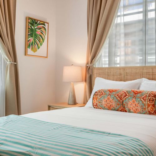 A bedroom with a queen-size bed dressed in white linens and a light blue throw. Two windows with beige curtains allow natural light. A wooden nightstand and soft lighting frame the bed.