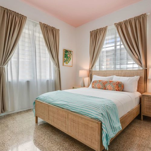 A bedroom with a queen-size bed dressed in white linens and a light blue throw. Two windows with beige curtains allow natural light. A wooden nightstand and soft lighting frame the bed.