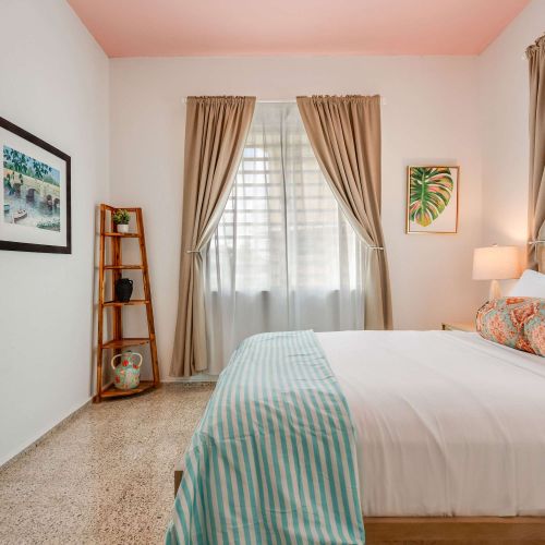 A bedroom with a queen-size bed dressed in white linens and a light blue throw. Two windows with beige curtains allow natural light. A wooden nightstand and soft lighting frame the bed.