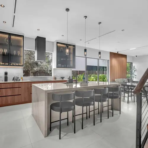 This high-end culinary space features a stunning contrast between warm wood lower cabinets and minimalist white floor-to-ceiling storage, anchored by a massive marble-style waterfall island.