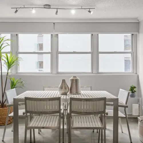 Second dining area with rectangular white table and six chairs beneath a row of windows with roller shades, potted palm and abstract art.