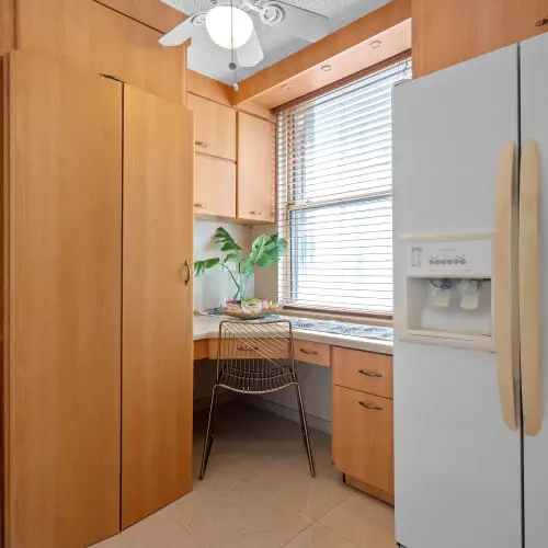 Built-in kitchen desk with gold wire chair beside a window with blinds, next to tall wood cabinets and the side-by-side refrigerator.