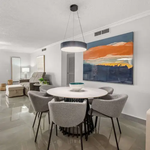 Dining area with round white table and six gray upholstered chairs under a black ring pendant, open to the wood-cabinet kitchen and the living room beyond.