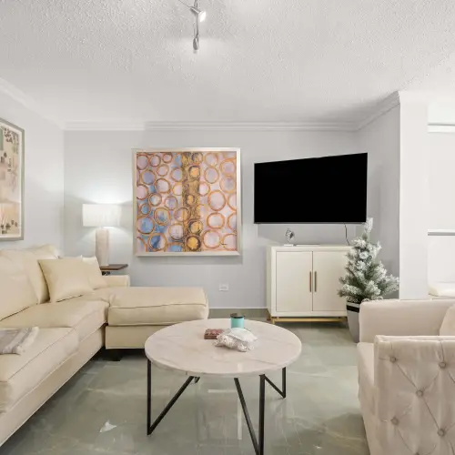 Living room with cream leather sectional, tufted swivel chair, round marble coffee table, wall-mounted TV on a white cabinet and track lighting.