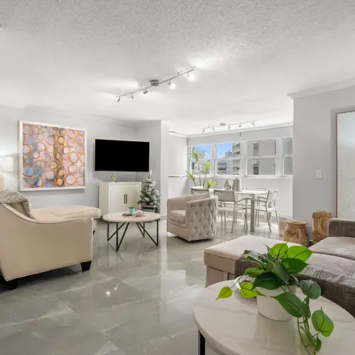Wide view of the living room with gray recliner sofa, cream sectional, marble side and coffee tables, and dining area under a row of windows.
