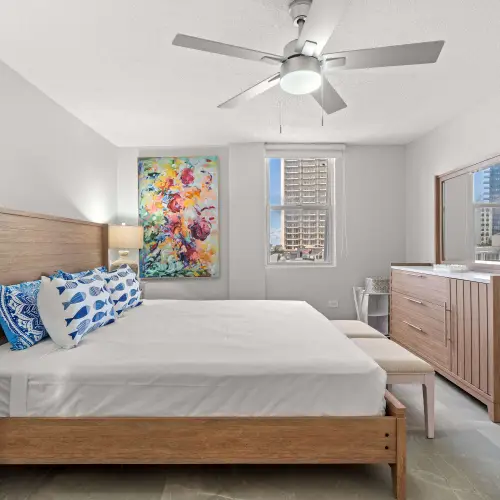 King bed with wood frame and fish-print pillows, floral canvas above, ceiling fan and wood dresser with mirror; city view through the window.