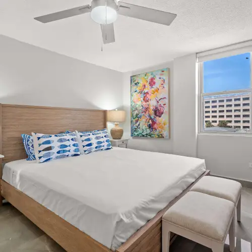 King bedroom with upholstered bench, woven-base lamps on white nightstands, colorful floral painting and window with city view.