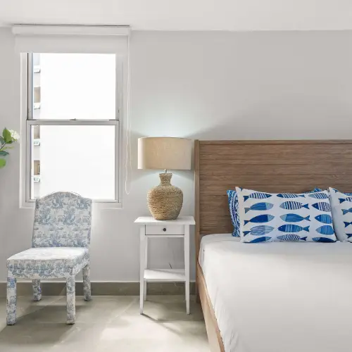 Corner of the king bedroom with blue toile chair, white nightstand with woven lamp, decorative flowering tree and window.