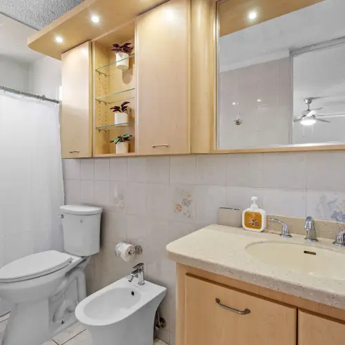 Bathroom with wood vanity, wide mirror, lit glass display shelves, toilet, bidet and tub with white shower curtain.