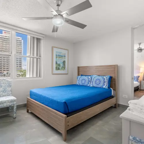 Bedroom with queen bed in a wood frame, blue bedspread, toile accent chair, ceiling fan and window with city views.