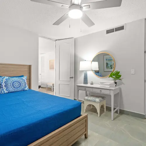 Queen bedroom with blue bedding, white vanity with stool and round mirror, ceiling fan, opening to the living area.