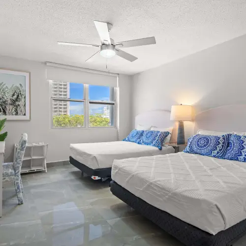 Bedroom with two queen beds, upholstered headboards and blue medallion pillows, desk with chair, palm print and window with greenery view.