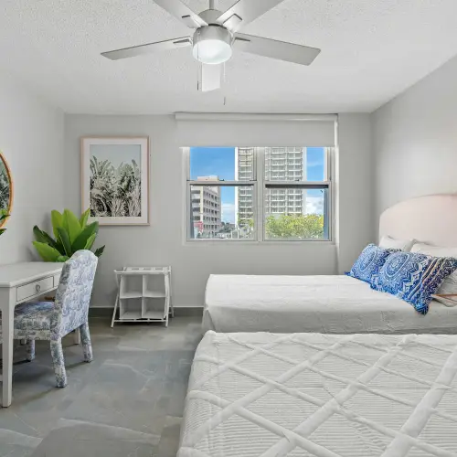 Two-queen bedroom with desk, round mirror, toile chair, luggage rack, ceiling fan and window facing city buildings.