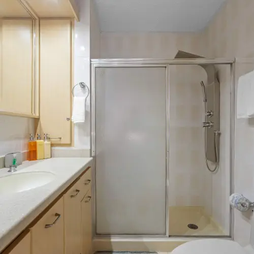 Bathroom with walk-in shower behind glass door, shower panel with handheld wand, wood vanity, wide mirror and fresh towels.