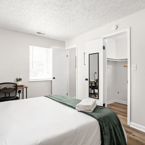 Experience a cozy, comfortable bed with soft pillows, complemented by a mirror, closet, work table, and chair with a desk lamp for your convenience and relaxation.