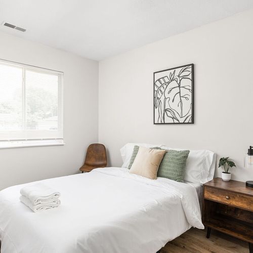 We've crafted a cozy space with a bed adorned in soft pillows and fresh linens. Bedside tables with lamps and a few frames on the wall enhance the ambiance, while a window adds to the inviting atmosphere.