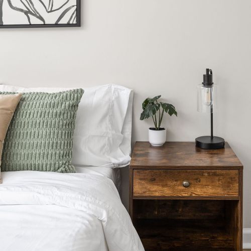Indulge in the comfort and style of soft pillows, framed wall art, a bedside table, a desk lamp, and a green plant in a white vase, all enhancing the ambiance of your space.