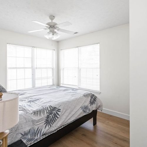 This cozy and inviting room adorned with two large windows, a bedside table, and a comforting lamp.
