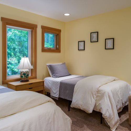 This bright bedroom features two comfortable twin beds, a shared nightstand with a reading lamp, a cozy accent chair, and large windows that fill the room with natural light and peaceful woodland views.