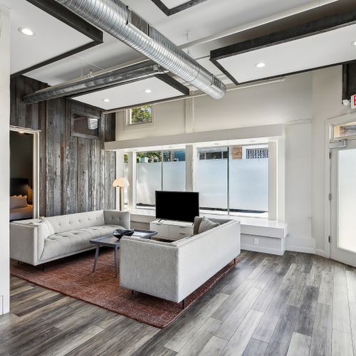 Bright living room w/ cozy sofa seating & a TV, framed by big windows.
