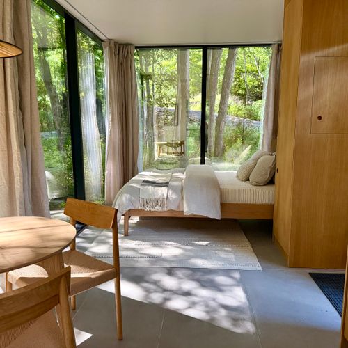 Open-plan guest studio: bed, dining, and forest views under one glass-wrapped roof.
