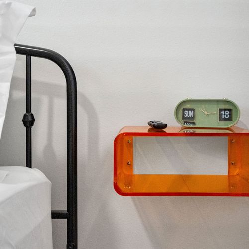 Bright orange floating nightstand & retro flip clock add a playful pop beside the classic iron bed